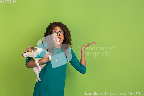 Image of African-american beautiful young woman\'s portrait with little doggy on green studio background, emotional and expressive. Copyspace for ad.