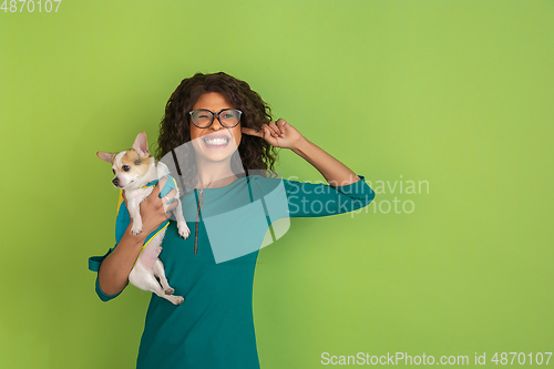 Image of African-american beautiful young woman\'s portrait with little doggy on green studio background, emotional and expressive. Copyspace for ad.