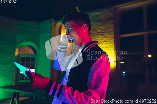 Image of Cinematic portrait of handsome young man using devices, gadgets in neon lighted interior. Youth culture, bright colors