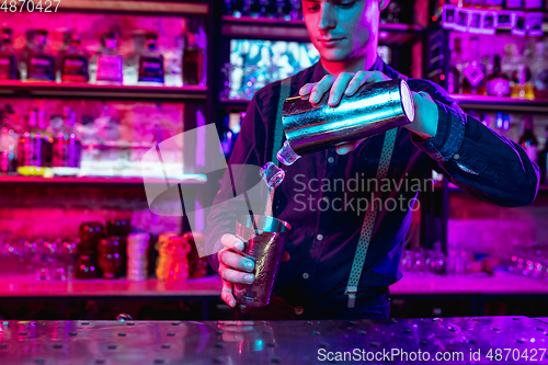 Image of Close up of barman finishes preparation of alcoholic cocktail with shaker in multicolored neon light