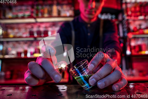 Image of Barman finishes preparation of alcoholic cocktail, sets fire to drink in multicolored neon light, focus on glass