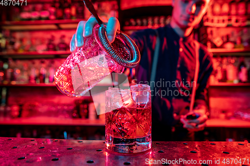 Image of Barman finishes preparation of alcoholic cocktail, pouring drink in multicolored neon light, focus on glass
