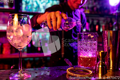Image of Close up of barman finishes preparation of alcoholic cocktail, pouring drink in multicolored neon light