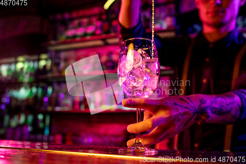 Image of Close up of barman finishes preparation of alcoholic cocktail, pouring drink in multicolored neon light