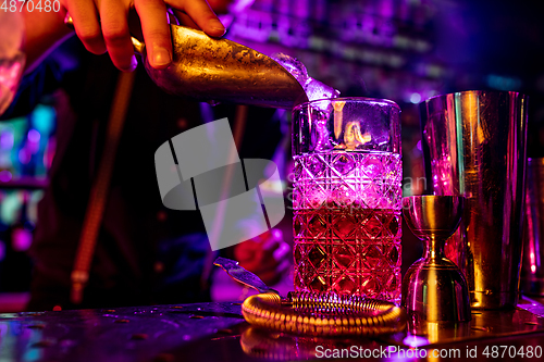 Image of Close up of barman finishes preparation of alcoholic cocktail, pouring drink in multicolored neon light