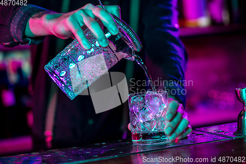 Image of Close up of barman finishes preparation of alcoholic cocktail, pouring drink in multicolored neon light