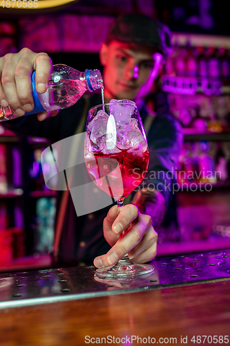 Image of Close up of barman finishes preparation of alcoholic cocktail, pouring drink in multicolored neon light, focus on glass