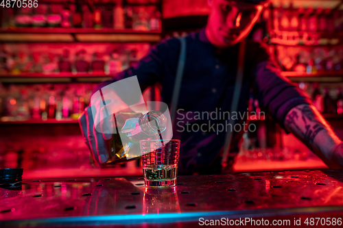 Image of Barman finishes preparation of alcoholic cocktail, pouring drink in multicolored neon light, focus on glass