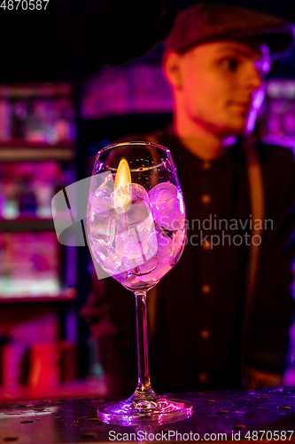 Image of Barman finishes preparation of alcoholic cocktail in multicolored neon light, focus on glass