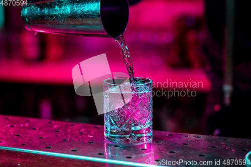 Image of Close up of barman finishes preparation of alcoholic cocktail, pouring drink in multicolored neon light