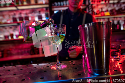 Image of Barman finishes preparation of alcoholic cocktail, pouring drink in multicolored neon light, focus on glass