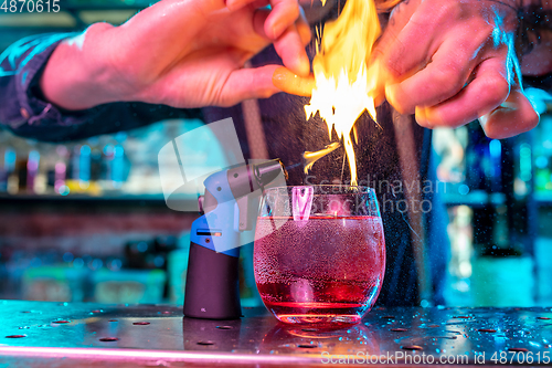 Image of Close up of alcoholic cocktail, beverage, drink in multicolored neon light with fire flames setting on orange\'s spraying juice