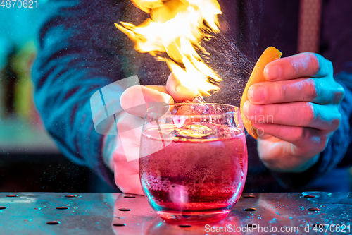 Image of Close up of alcoholic cocktail, beverage, drink in multicolored neon light with fire flames setting on orange\'s spraying juice