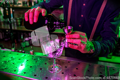 Image of Close up of barman finishes preparation of alcoholic cocktail, pouring drink with shot in multicolored neon light, focus on glass