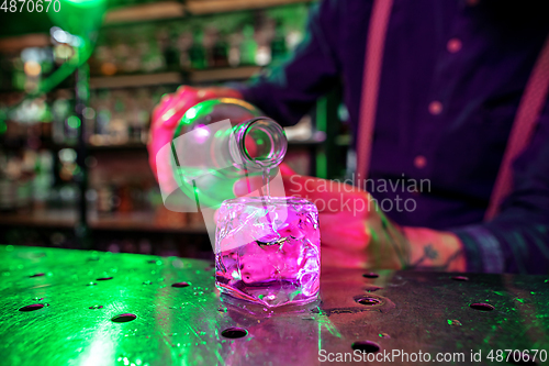 Image of Close up of barman finishes preparation of alcoholic cocktail, pouring drink in multicolored neon light, focus on glass