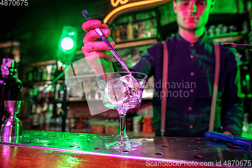 Image of Close up of barman finishes preparation of alcoholic cocktail, pouring drink in multicolored neon light, focus on glass