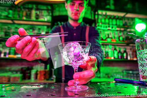 Image of Close up of barman finishes preparation of alcoholic cocktail, pouring drink in multicolored neon light, focus on glass