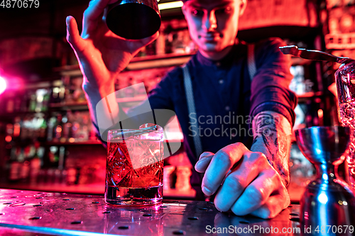 Image of Close up of barman finishes preparation of alcoholic cocktail, pouring drink in multicolored neon light, focus on glass