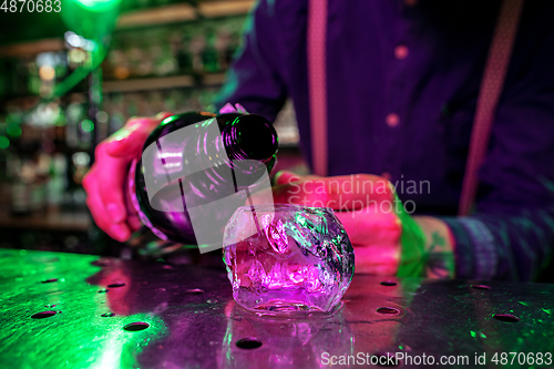 Image of Close up of barman finishes preparation of alcoholic cocktail, pouring drink in multicolored neon light, focus on glass