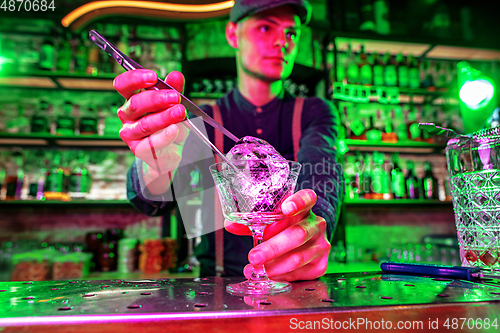 Image of Close up of barman finishes preparation of alcoholic cocktail, pouring drink in multicolored neon light, focus on glass