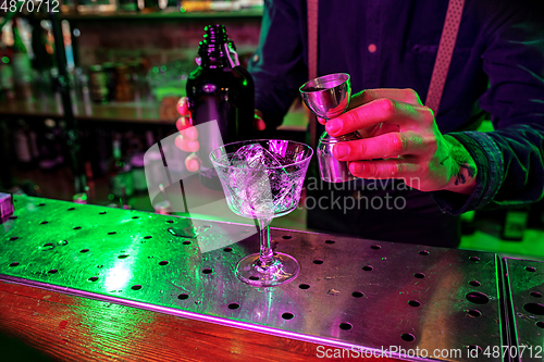 Image of Close up of barman finishes preparation of alcoholic cocktail, pouring drink with shot in multicolored neon light, focus on glass