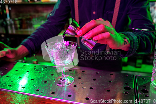 Image of Close up of barman finishes preparation of alcoholic cocktail, pouring drink with shot in multicolored neon light, focus on glass