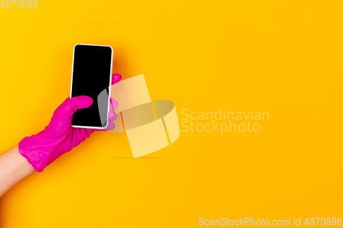 Image of Hand in pink rubber glove holding smartphone with blank screen isolated on yellow studio background with copyspace.