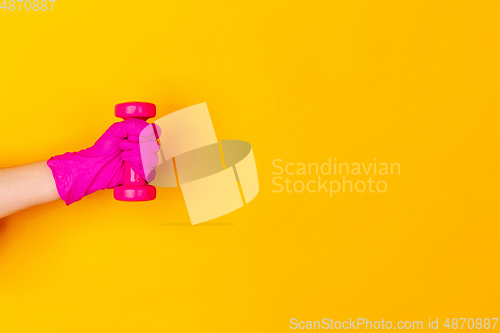 Image of Hand in pink rubber glove holding dumbbell, gym weight isolated on yellow studio background with copyspace.