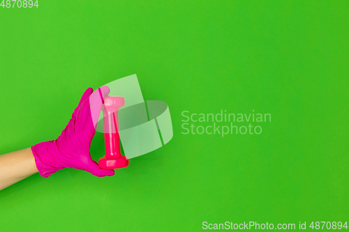 Image of Hand in pink rubber glove holding gym weight, dumbbell isolated on green studio background with copyspace.