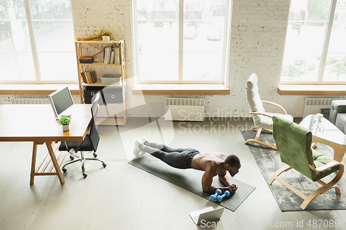 Image of Young african-american man training at home during quarantine of coronavirus outbreak, doinc exercises of fitness, aerobic. Staying sportive suring insulation.