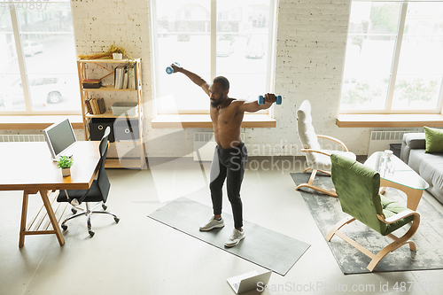 Image of Young african-american man training at home during quarantine of coronavirus outbreak, doinc exercises of fitness, aerobic. Staying sportive suring insulation.