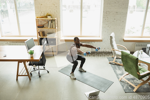 Image of Young african-american man training at home during quarantine of coronavirus outbreak, doinc exercises of fitness, aerobic. Staying sportive suring insulation.