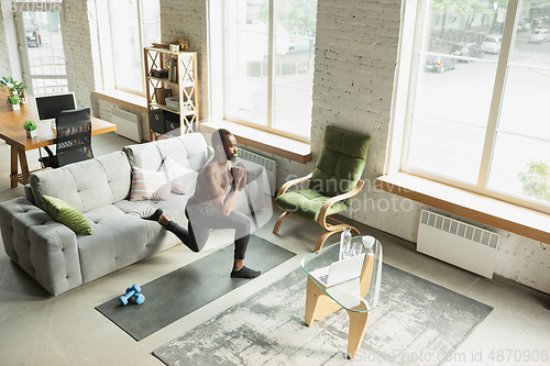 Image of Young african-american man training at home during quarantine of coronavirus outbreak, doinc exercises of fitness, aerobic. Staying sportive suring insulation.