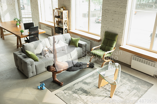 Image of Young african-american man training at home during quarantine of coronavirus outbreak, doinc exercises of fitness, aerobic. Staying sportive suring insulation.