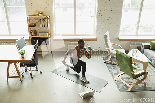 Image of Young african-american man training at home during quarantine of coronavirus outbreak, doinc exercises of fitness, aerobic. Staying sportive suring insulation.