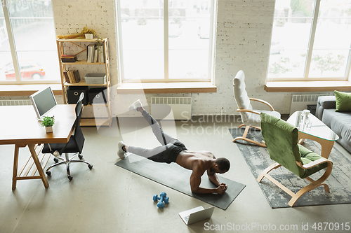 Image of Young african-american man training at home during quarantine of coronavirus outbreak, doinc exercises of fitness, aerobic. Staying sportive suring insulation.