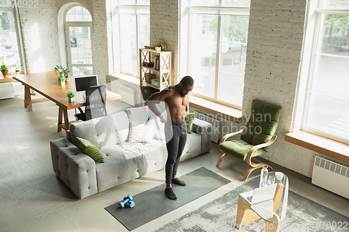 Image of Young african-american man training at home during quarantine of coronavirus outbreak, doinc exercises of fitness, aerobic. Staying sportive suring insulation.