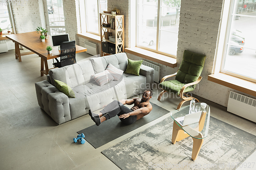 Image of Young african-american man training at home during quarantine of coronavirus outbreak, doinc exercises of fitness, aerobic. Staying sportive suring insulation.