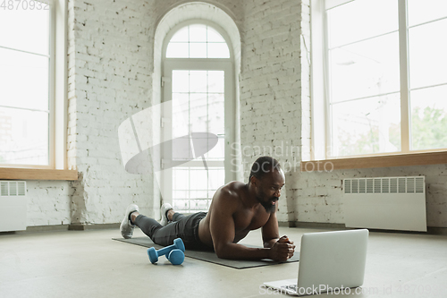 Image of Young african-american man training at home during quarantine of coronavirus outbreak, doinc exercises of fitness, aerobic. Staying sportive suring insulation.