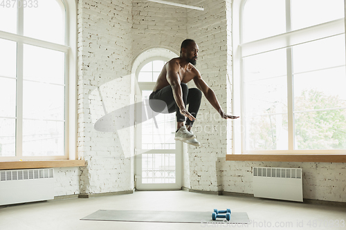 Image of Young african-american man training at home during quarantine of coronavirus outbreak, doinc exercises of fitness, aerobic. Staying sportive suring insulation.