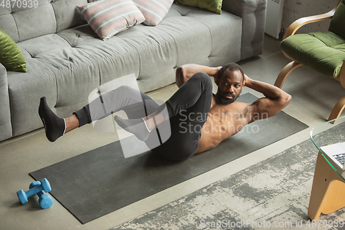 Image of Young african-american man training at home during quarantine of coronavirus outbreak, doinc exercises of fitness, aerobic. Staying sportive suring insulation.