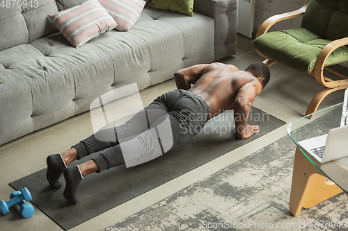 Image of Young african-american man training at home during quarantine of coronavirus outbreak, doinc exercises of fitness, aerobic. Staying sportive suring insulation.