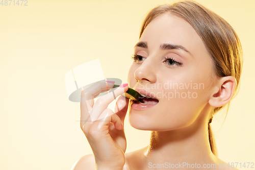 Image of Biting. Close up of beautiful female face with cucumber slice over yellow background. Cosmetics and makeup, natural and eco treatment, skin care.