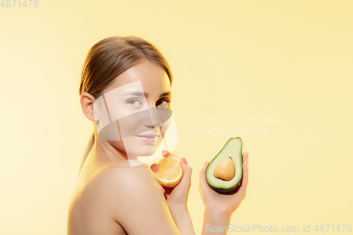 Image of Vitamins splash. Close up of beautiful female face with lemon and avocado halfs over yellow background. Cosmetics and makeup, natural and eco treatment, skin care.