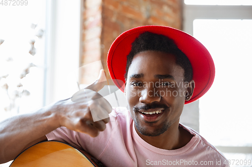 Image of African-american musician in red hat greeting audience before online concert at home isolated and quarantined, cheerful, smiling
