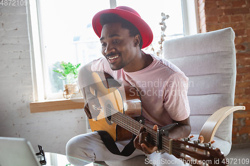 Image of African-american musician playing guitar during online concert at home isolated and quarantined, impressive improvising