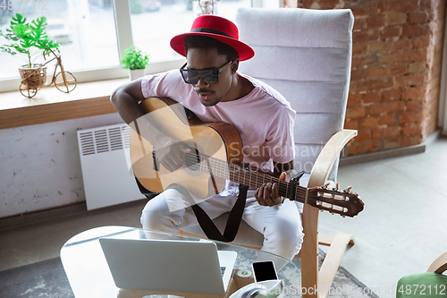 Image of African-american musician playing guitar during online concert at home isolated and quarantined, impressive improvising