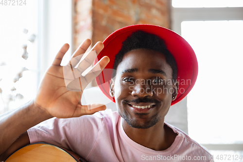 Image of African-american musician in red hat greeting audience before online concert at home isolated and quarantined, cheerful, smiling
