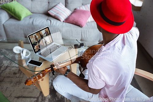 Image of African-american musician playing guitar during concert at home isolated and quarantined, impressive improvising with band connected online