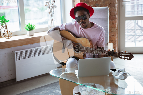 Image of African-american musician playing guitar during online concert at home isolated and quarantined, impressive improvising